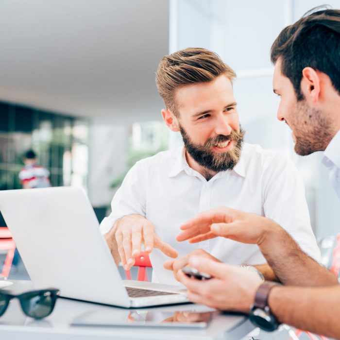 Two young bearded caucasian modern business man sitting in a bar, using laptop, looking each other, smiling and chatting - business, work, technology concept